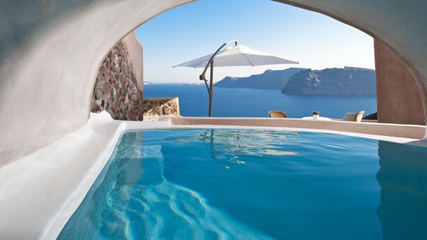 A clear view of a swimming pool with a stone wall and a white umbrella. - Armeni Luxury Villas (Santorini)