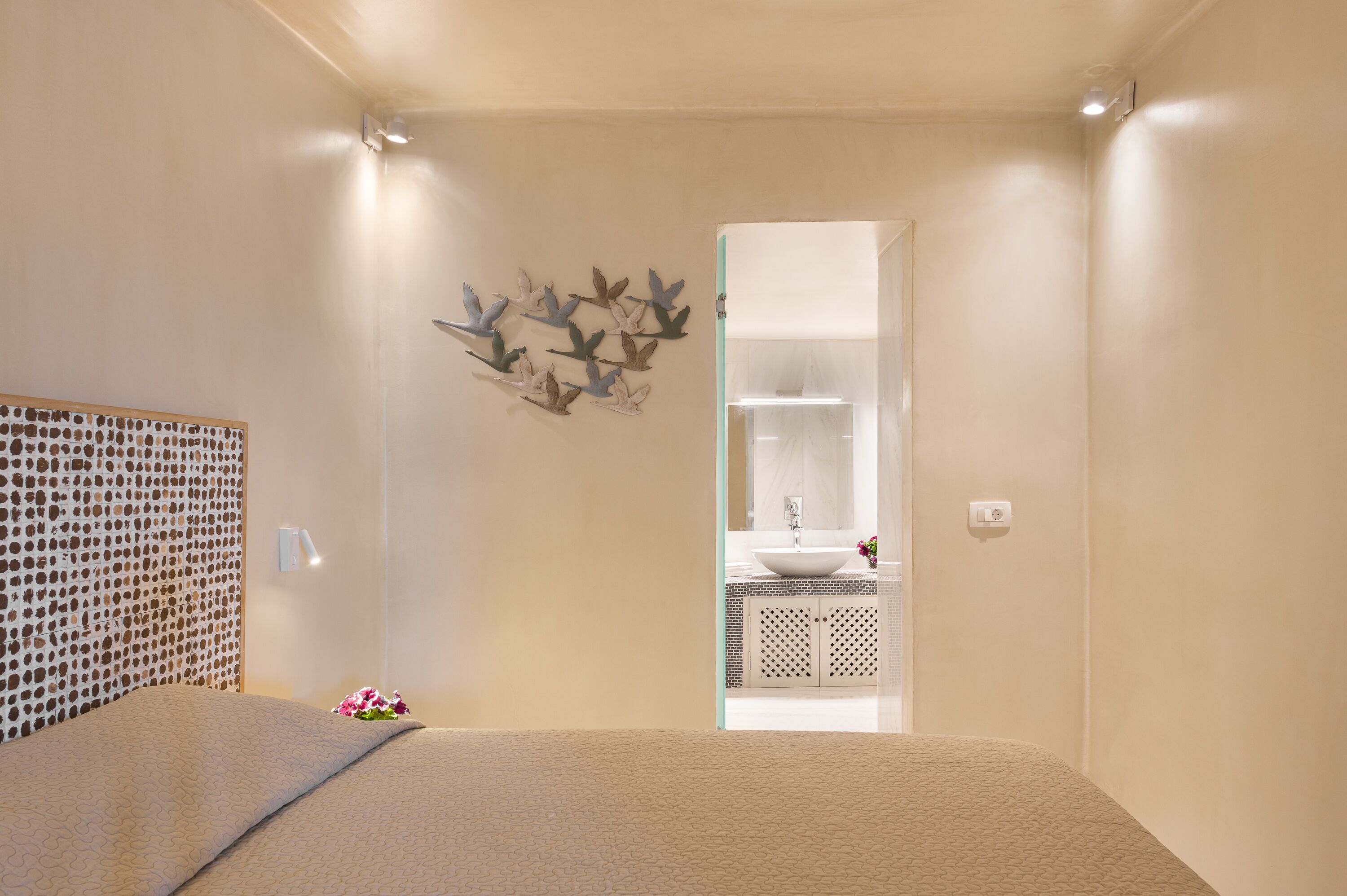 A bedroom with a bed, a bedside table, a wall-mounted light, a decorative wall piece, and a bathroom visible through an open door.