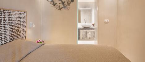 A bedroom with a bed, a bedside table, a wall-mounted light, a decorative wall piece, and a bathroom visible through an open door.