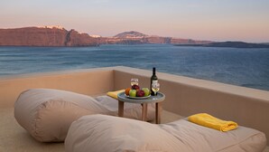 A balcony with a table set with fruits, a bottle, and glasses, overlooking a body of water and cliffs. - Armeni Luxury Villas (Santorini)