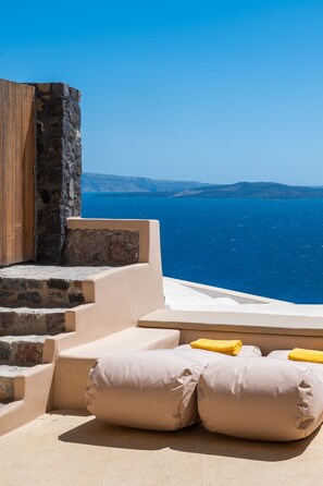 Un espacio exterior con mobiliario iluminado por el sol, cojines color beige y una pared de piedra, con vistas a un mar azul cristalino y la costa en la distancia.