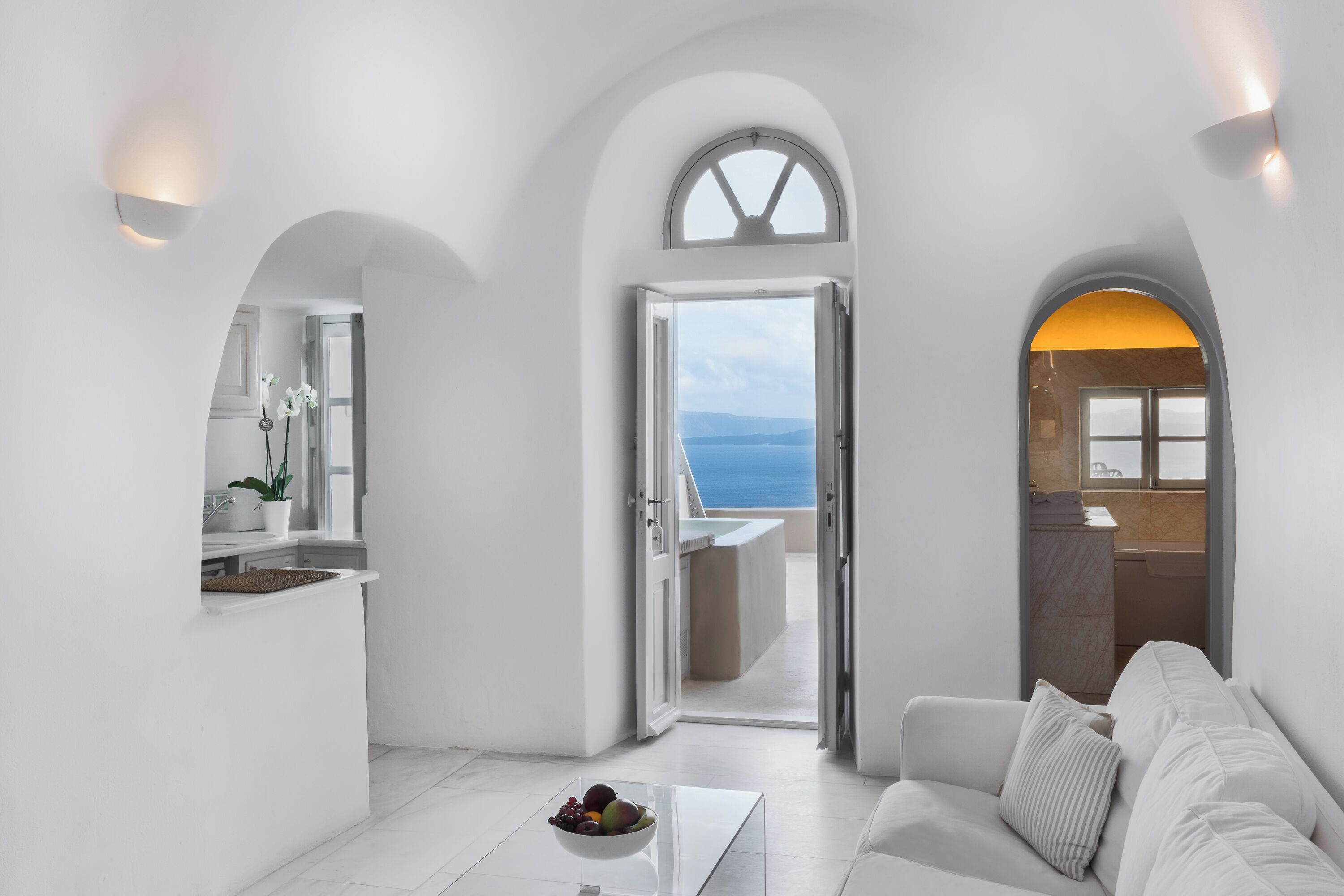 Un intérieur moderne aux couleurs claires, avec un coin repas agrémenté d’un saladier de fruits, un canapé et une vue sur la mer par une porte ouverte.