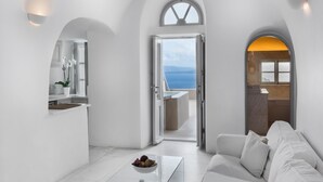 A modern, minimalist living room with a white sofa, a coffee table with fruit, and a view of the sea through an open door. - Armeni Luxury Villas (Santorini)