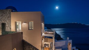 A coastal building with a stone facade, a balcony with a bed, and a view of the sea at night. - Armeni Luxury Villas (Santorini)