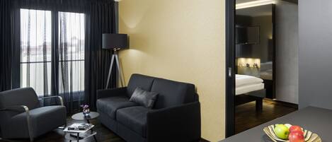 Una habitación de hotel moderna con un sofá de cuero oscuro, una pequeña mesa redonda de centro, un televisor de pantalla plana y una mesa de comedor con fruta.