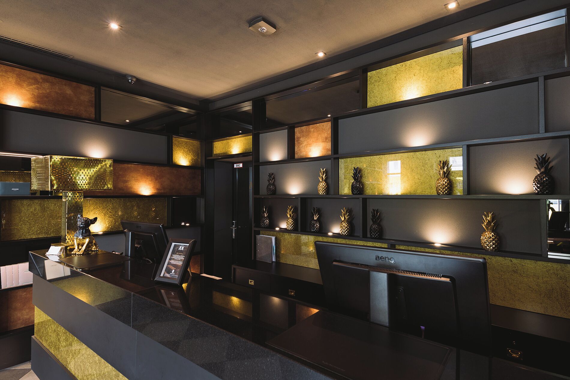 A modern hotel lobby with a reception desk, a digital information board, and a wall with decorative pineapple ornaments.