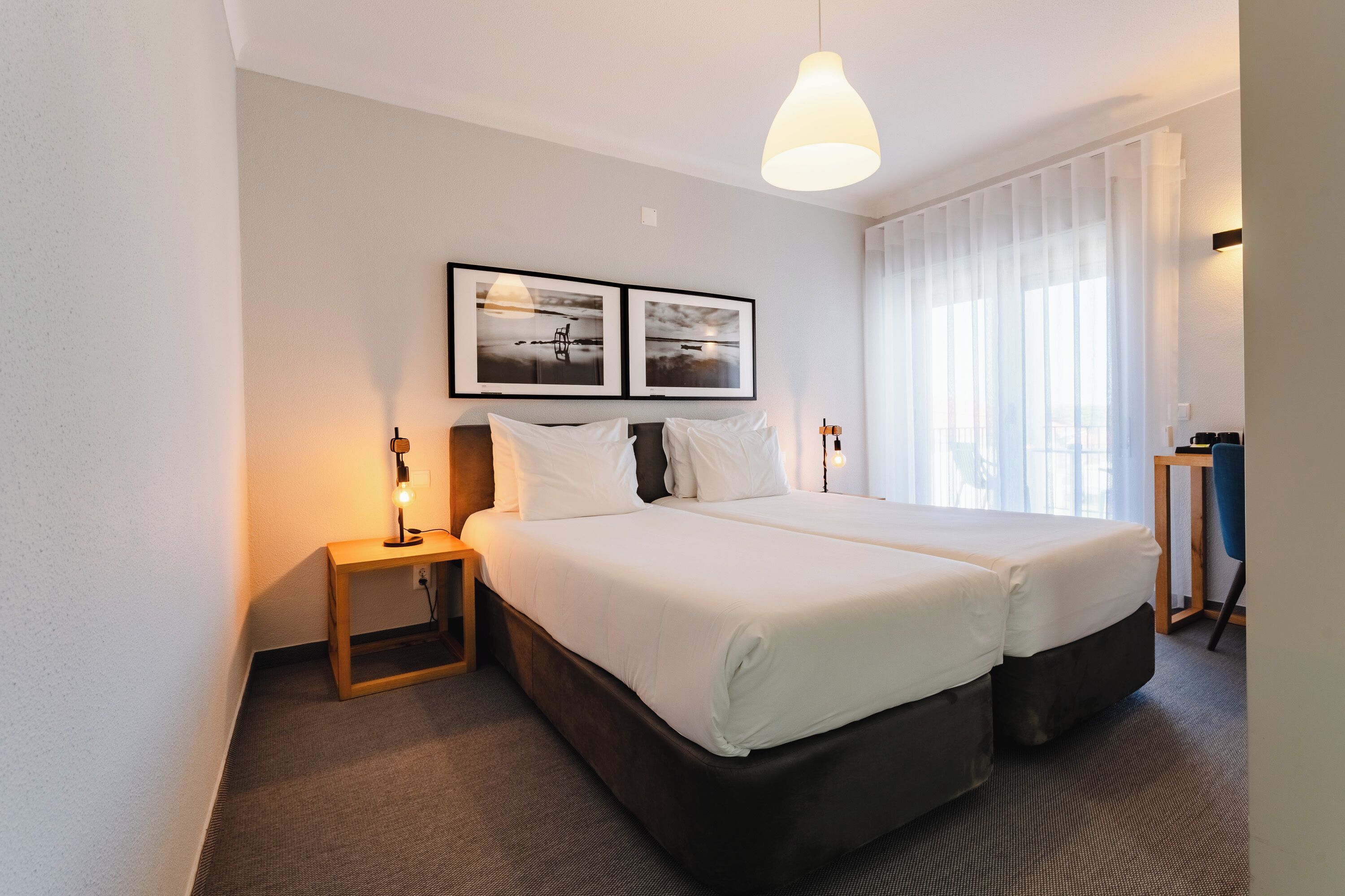 A hotel room with a double bed, a bedside table with a lamp, a large window with sheer curtains, and two framed pictures on the wall.