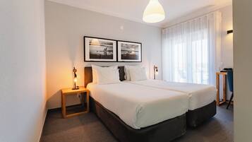 A hotel room with a double bed, a bedside table with a lamp, a large window with sheer curtains, and two framed pictures on the wall.