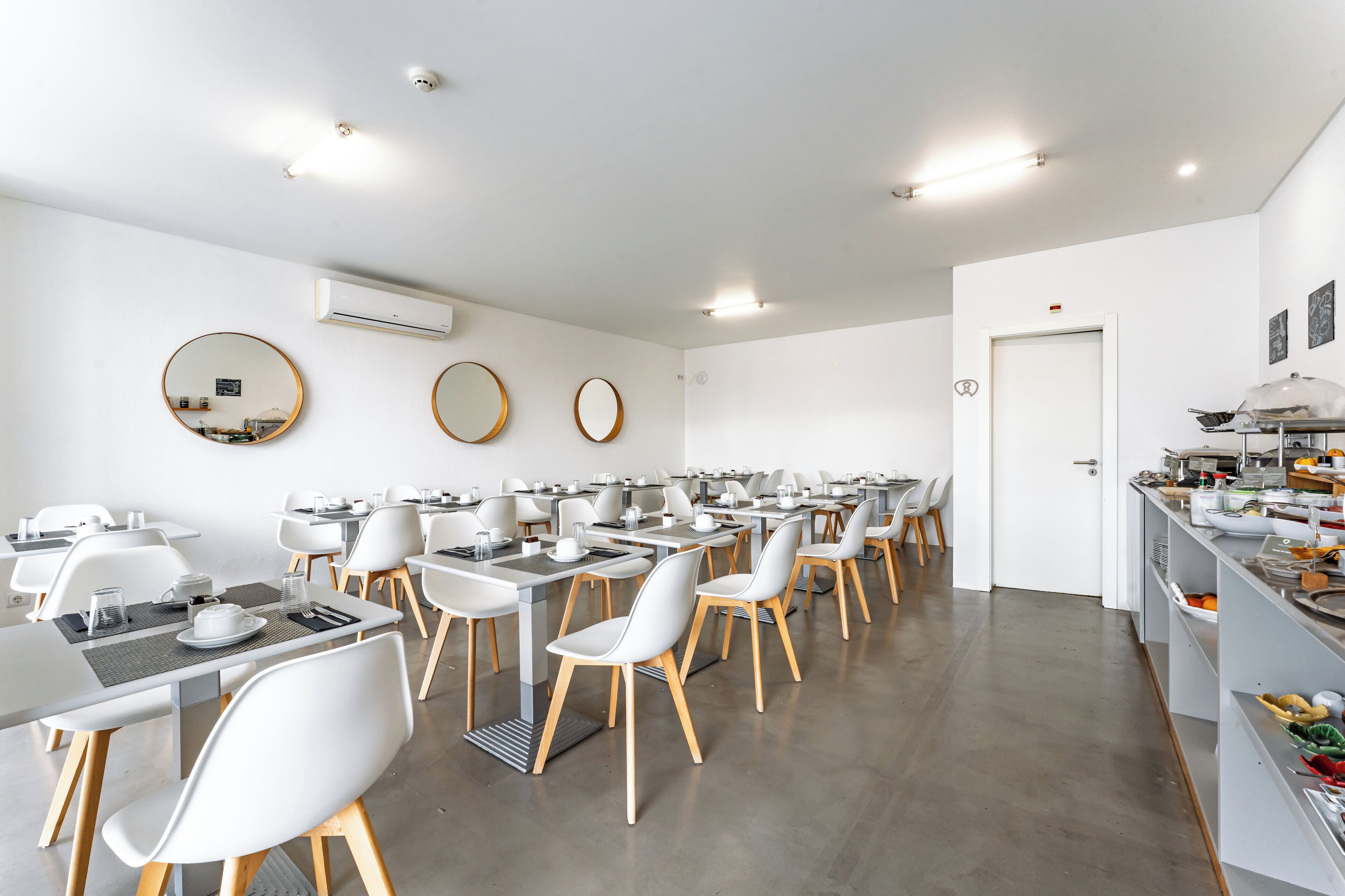 Un comedor moderno con mesas y sillas blancas, un mostrador tipo buffet y una puerta blanca.