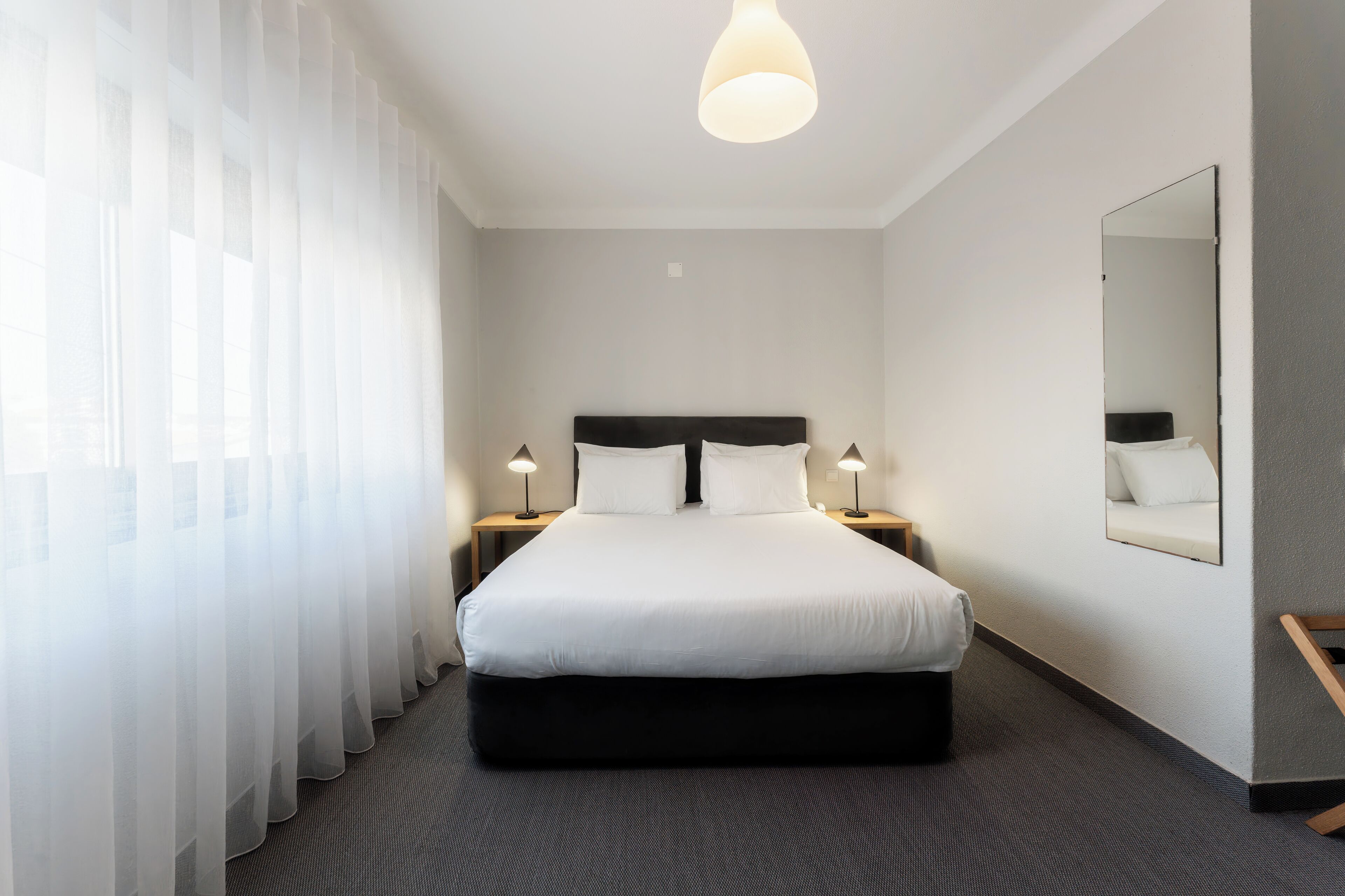 Una habitación de hotel con una cama grande, dos mesitas de noche con lámparas, un espejo en la pared y cortinas traslúcidas.