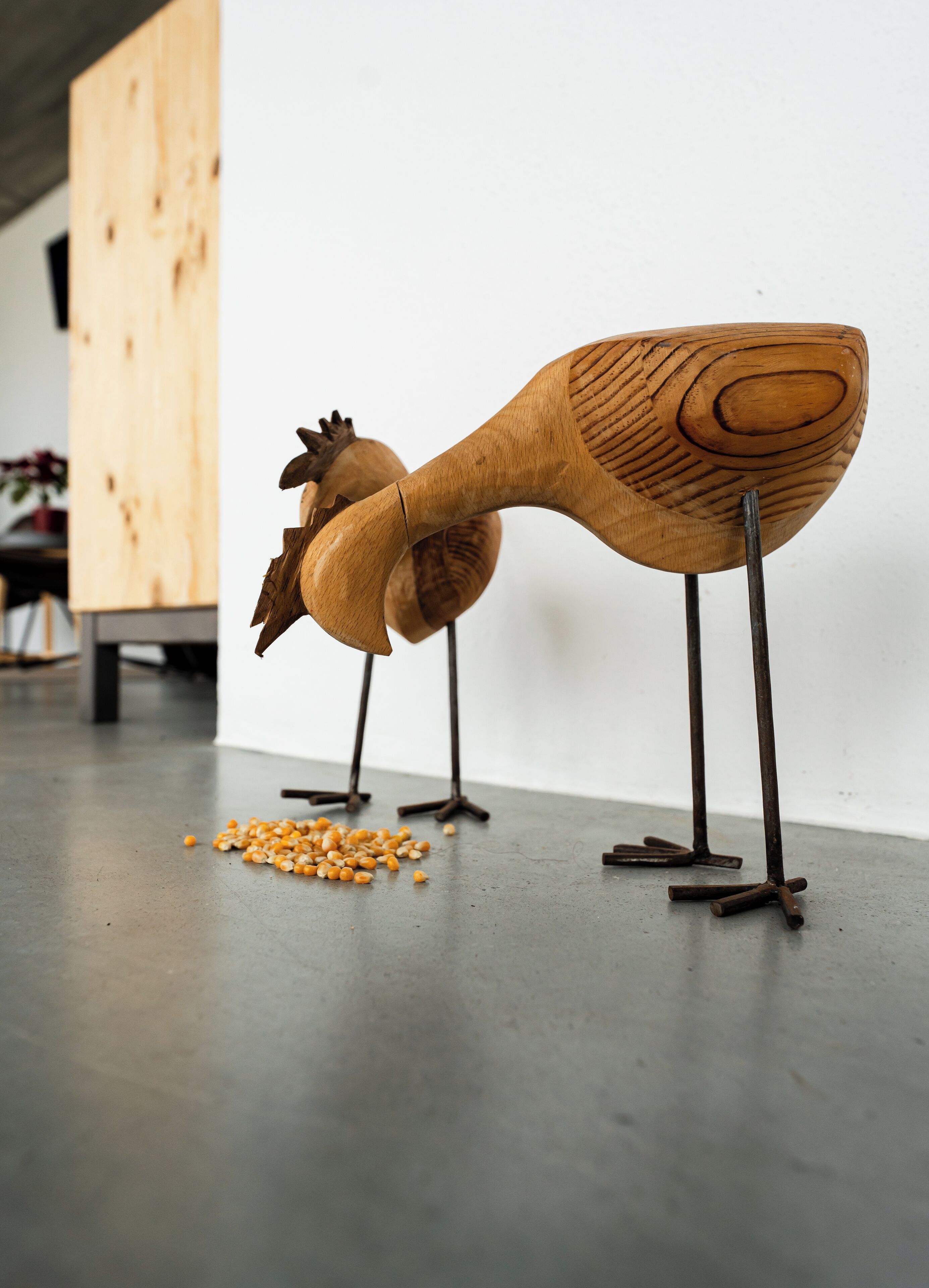Dos esculturas de madera de aves sobre soportes metálicos en una habitación con suelo de concreto y un armario de madera al fondo.