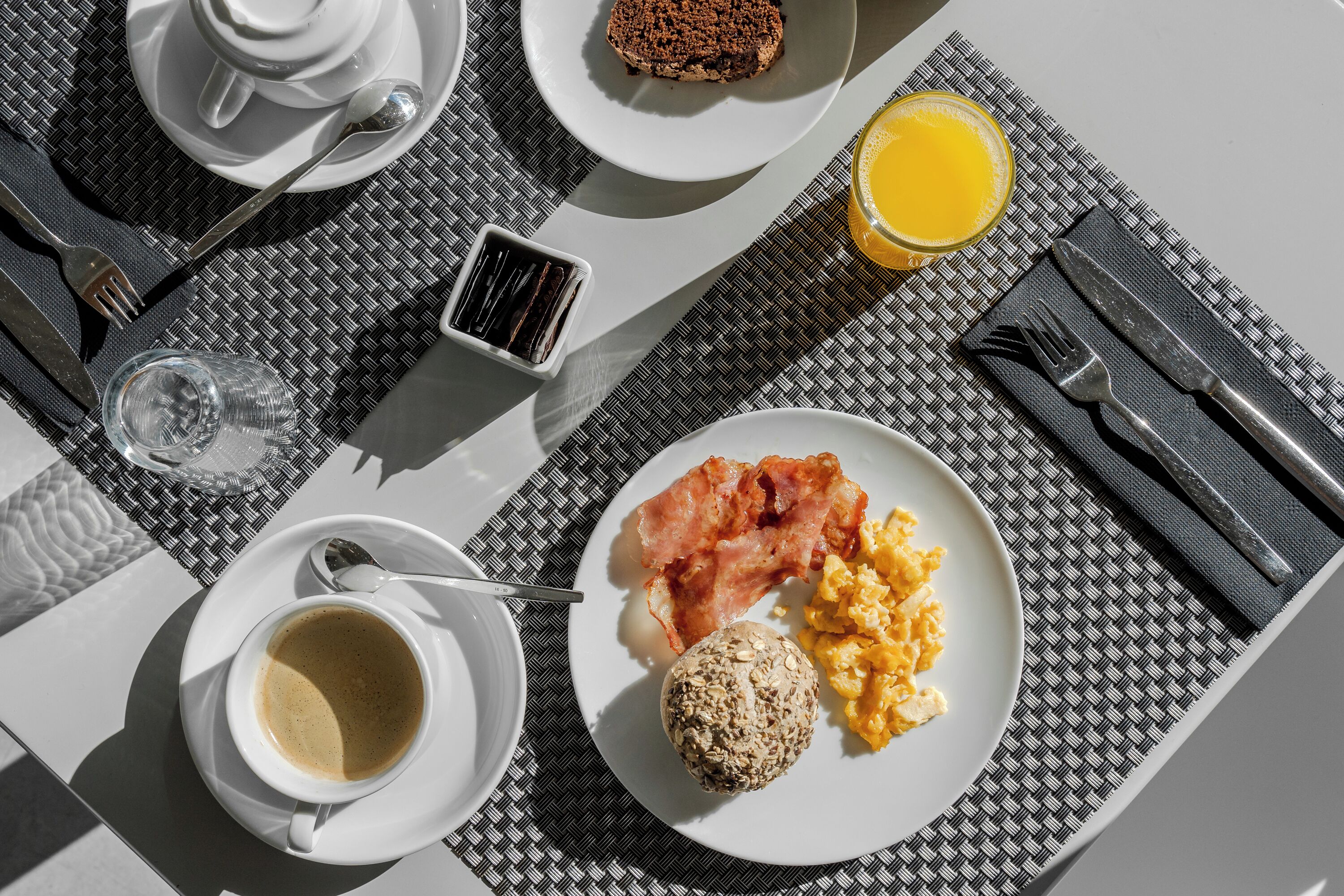 A breakfast setting with a plate of scrambled eggs, bacon, and a muffin, a cup of coffee, and a glass of orange juice on a table with a black and white checkered placemat.