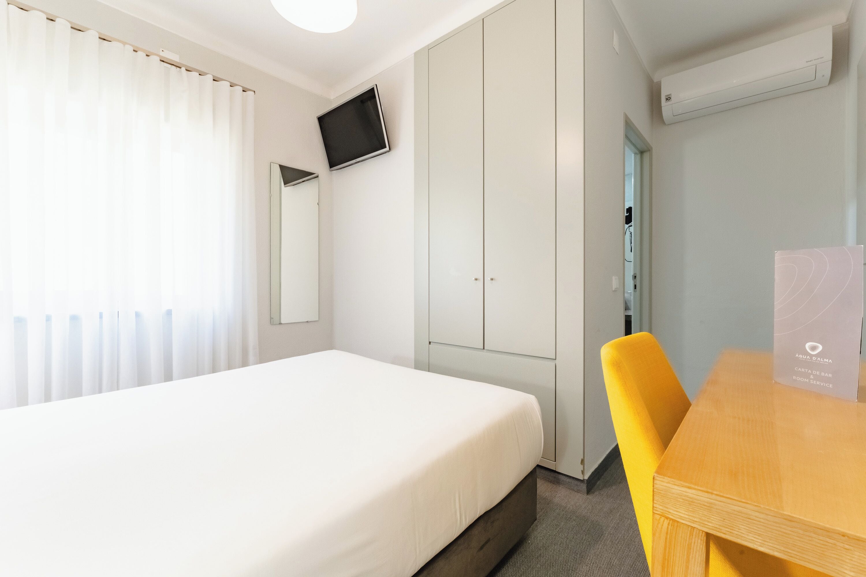 Una habitación de hotel con cama, escritorio, silla, televisor montado en la pared y armario.