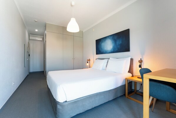 Una habitación de hotel con una cama, un escritorio, una silla, un cuadro en la pared y una lámpara en el techo.