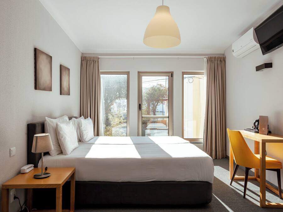 Una camera d'albergo con un letto, una scrivania, una sedia, una lampada, un televisore e un condizionatore d'aria.