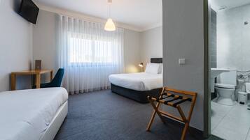 Quarto de hotel com duas camas, uma escrivaninha, uma cadeira, televisão, janela com cortinas e banheiro com vaso sanitário e pia.