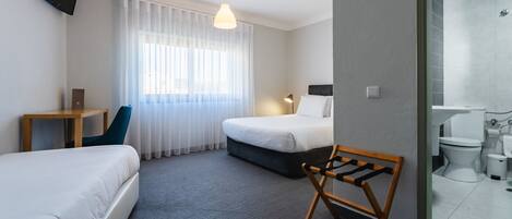 A hotel room with two beds, a desk, a chair, a TV, a window with curtains, and a bathroom with a toilet and sink.