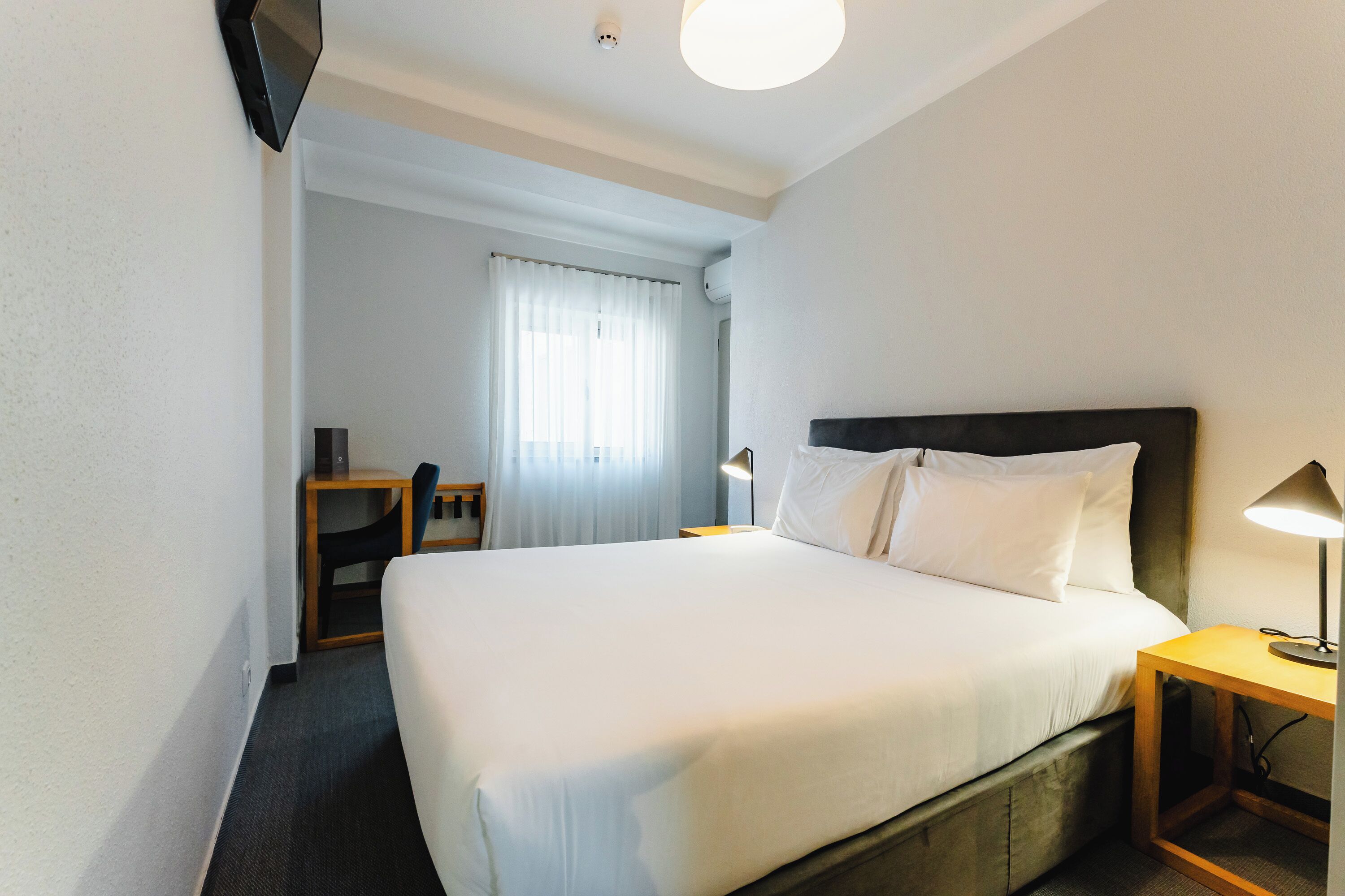 A hotel room with a large bed, a desk, a chair, a lamp, and a TV mounted on the wall.