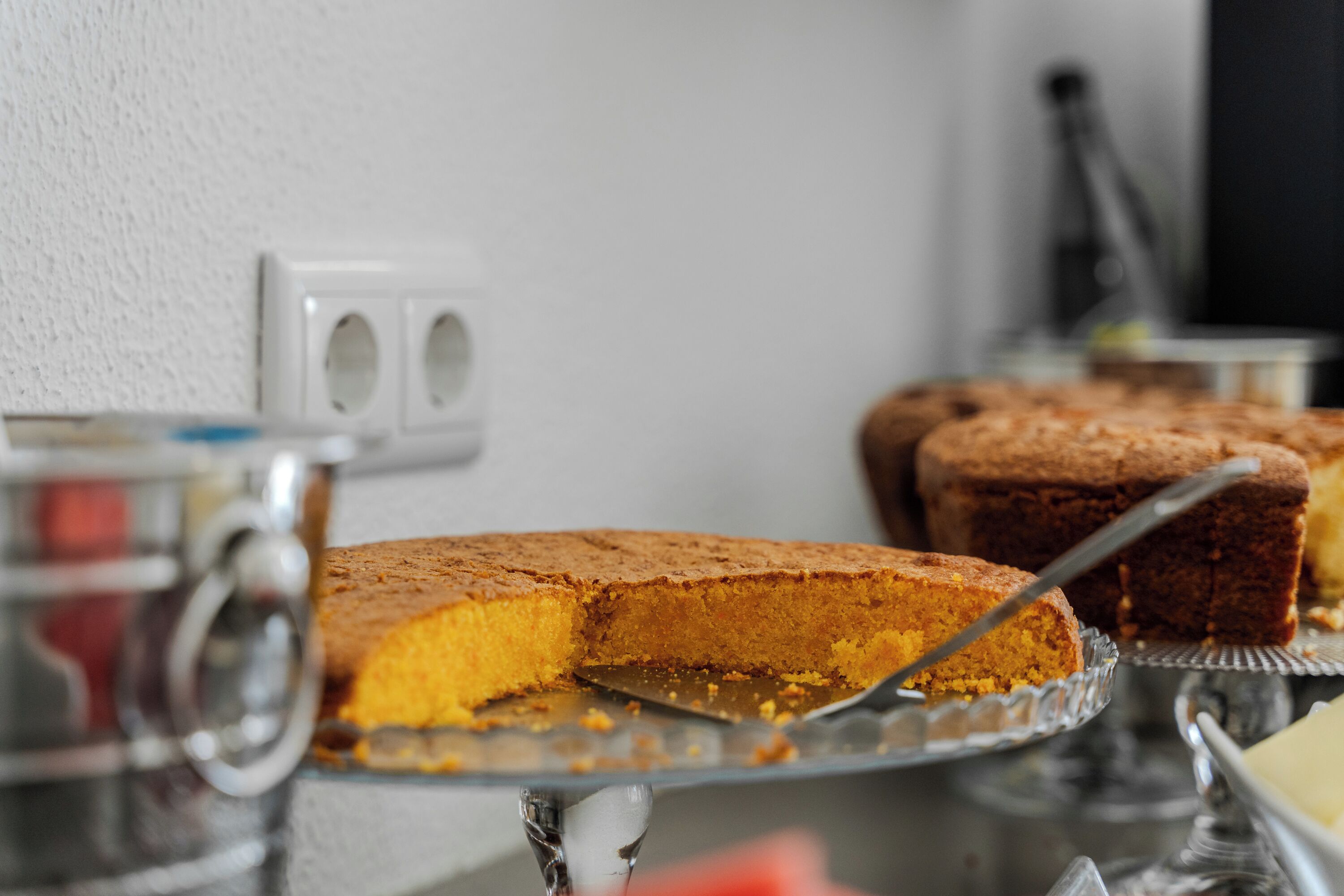 A cake on a glass stand with a knife, a white wall with electrical outlets, and a blurred background.
