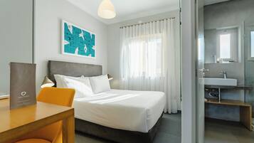 Une chambre d’hôtel comprenant un lit, un bureau, une chaise, une table de chevet, une lampe, une fenêtre avec des rideaux, une salle de bain avec un lavabo et un miroir, et une œuvre d’art accrochée au mur.
