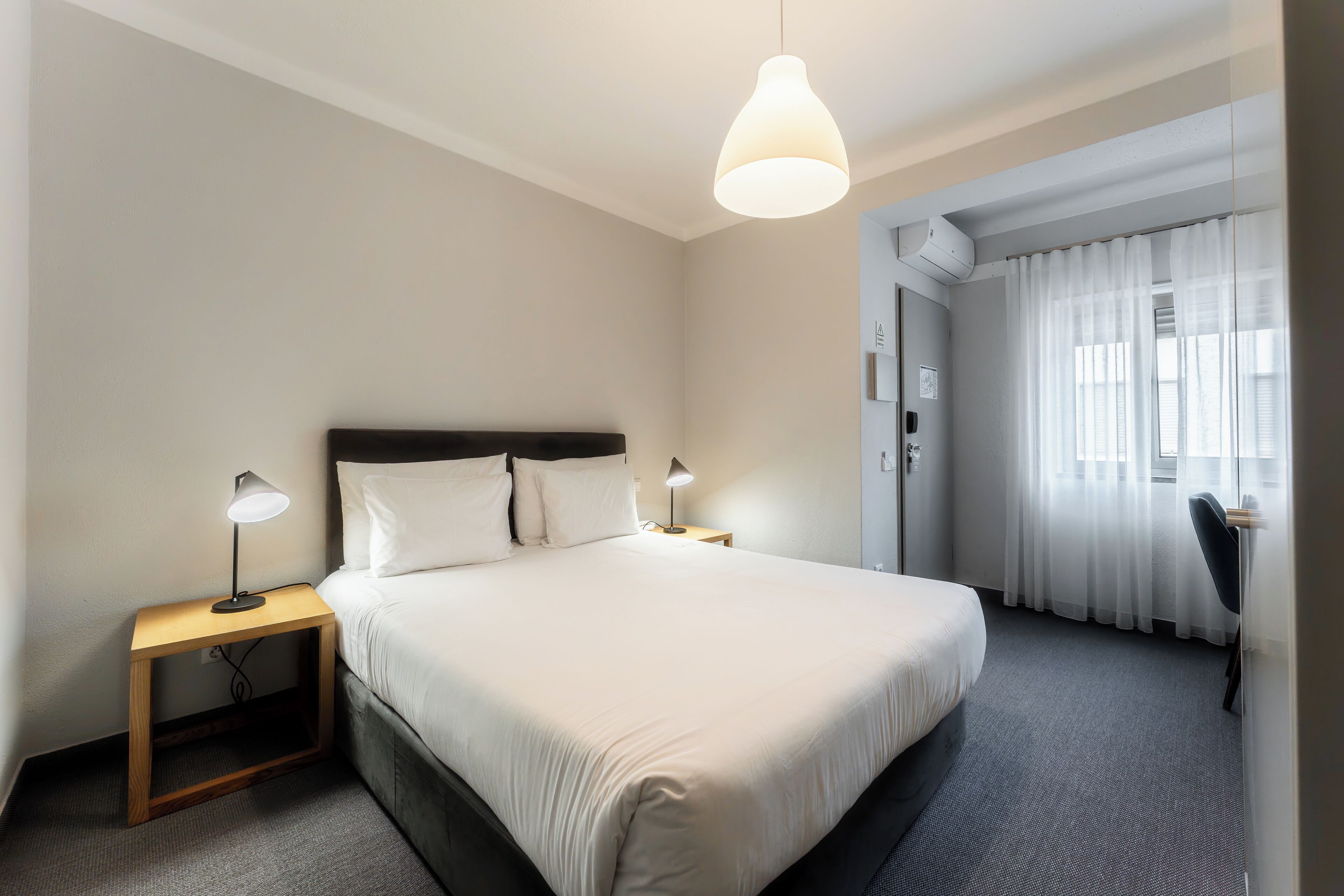 A hotel room with a large bed, two bedside tables with lamps, a desk with a chair, and a window with sheer curtains.