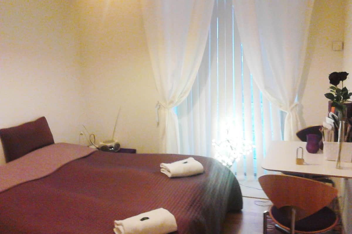 Quarto de hotel com cama, escrivaninha, cadeira, luminária e janela com cortinas.