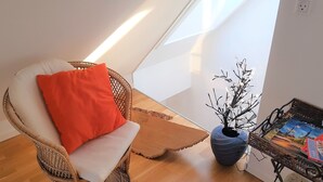 Une pièce avec un fauteuil en osier, une petite table sur laquelle est posé un vase rempli de branches, et une vue sur le ciel grâce à une lucarne.