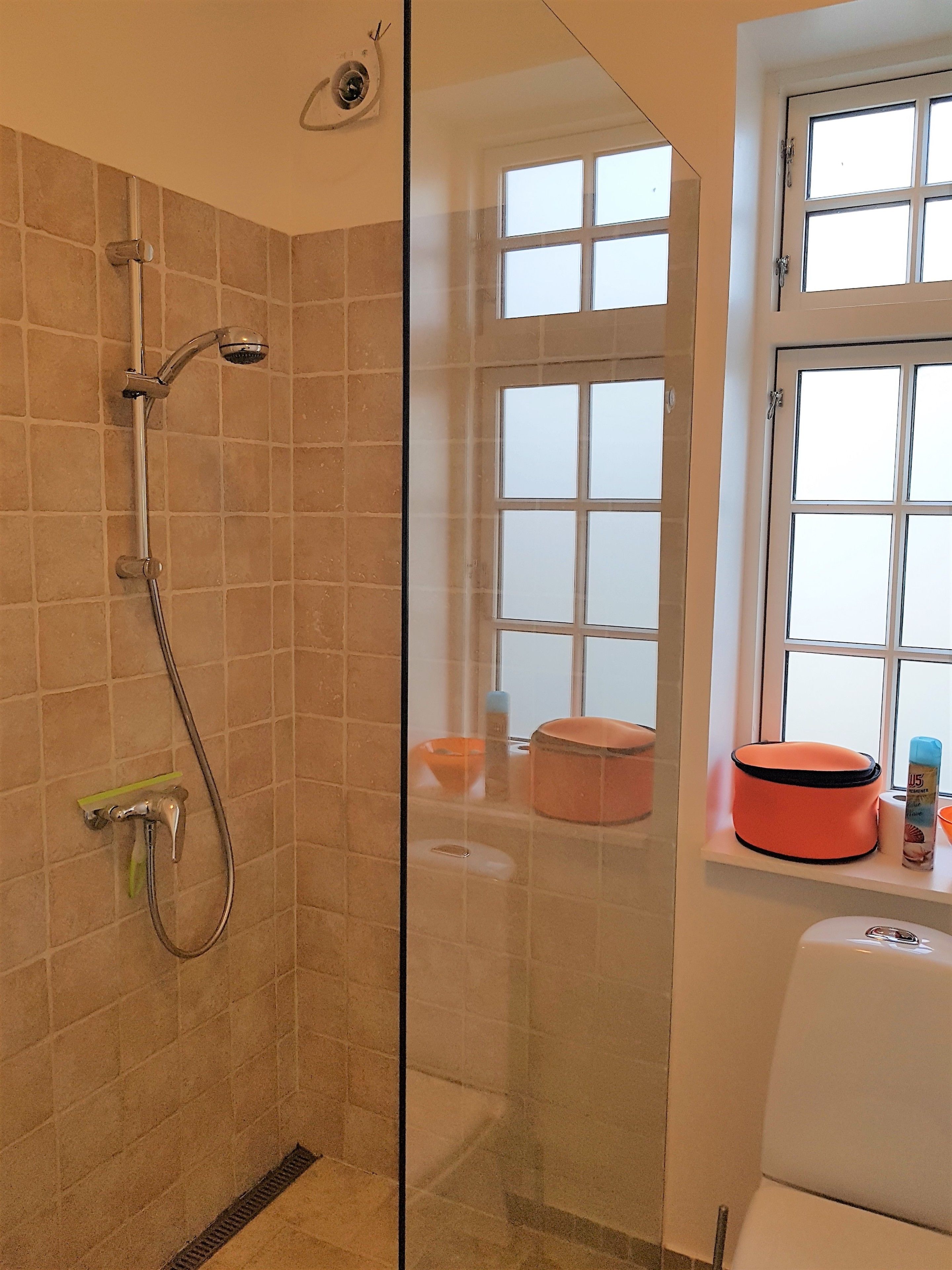 Ducha con puerta de vidrio, ventana con persianas y lavamanos con artículos de tocador.