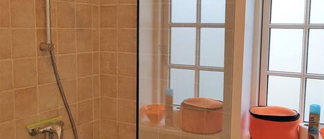 A shower with a glass door, a window with blinds, and a sink with toiletries.