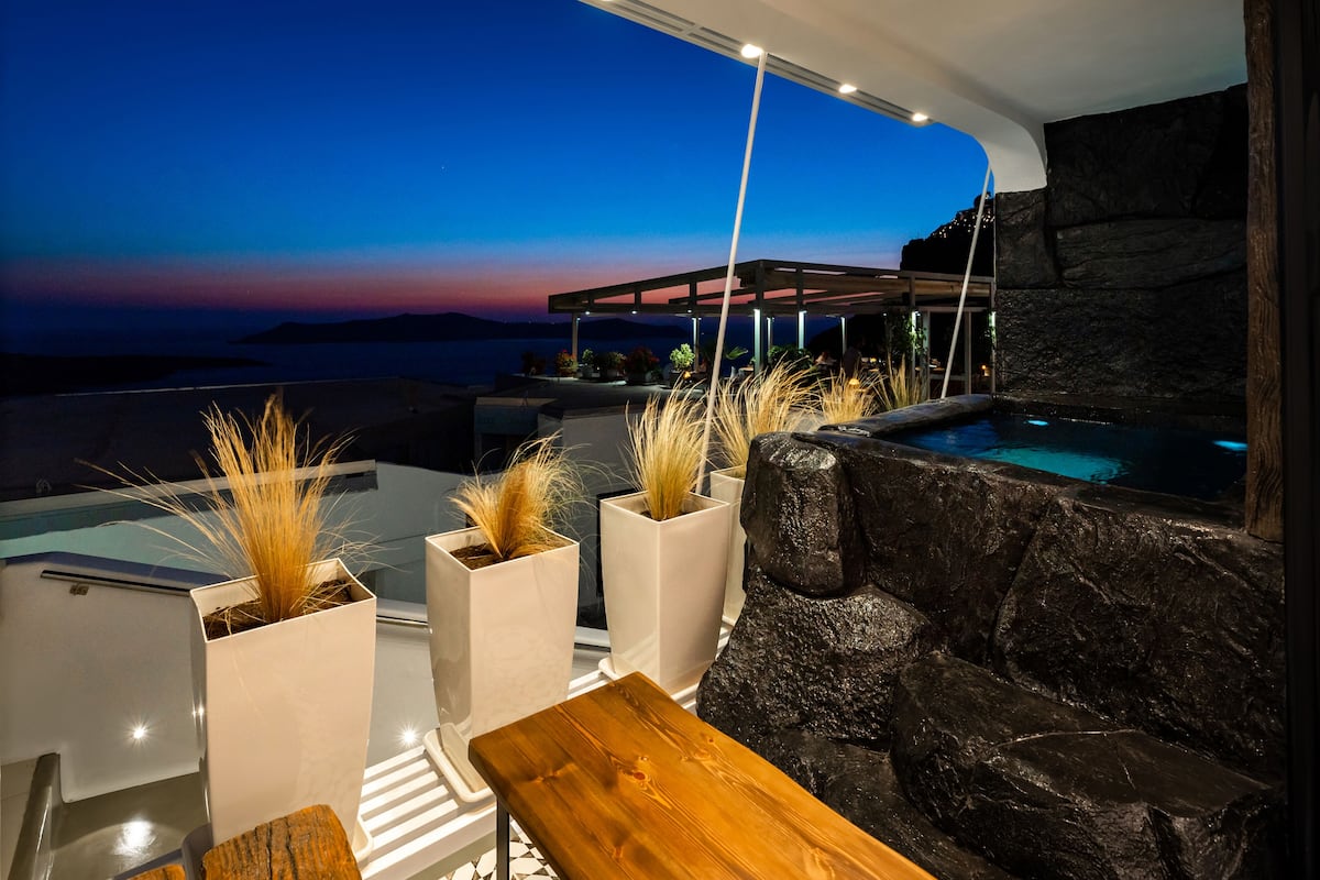 A modern outdoor seating area with a view of the sea at dusk, featuring a stone wall, a wooden bench, and potted plants.