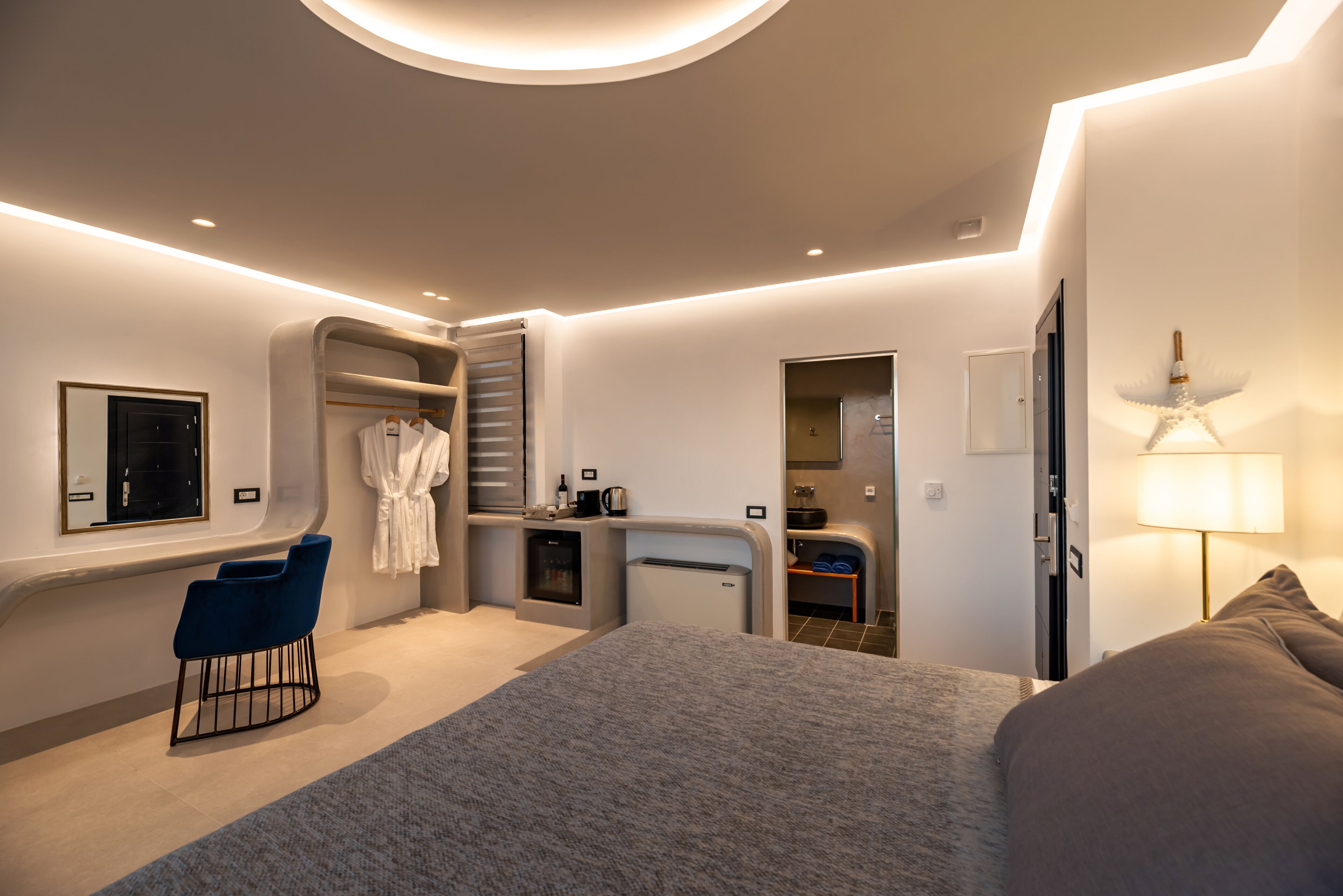 A modern hotel room with a large bed, a seating area with a chair, a vanity with a mirror, and a small kitchenette.