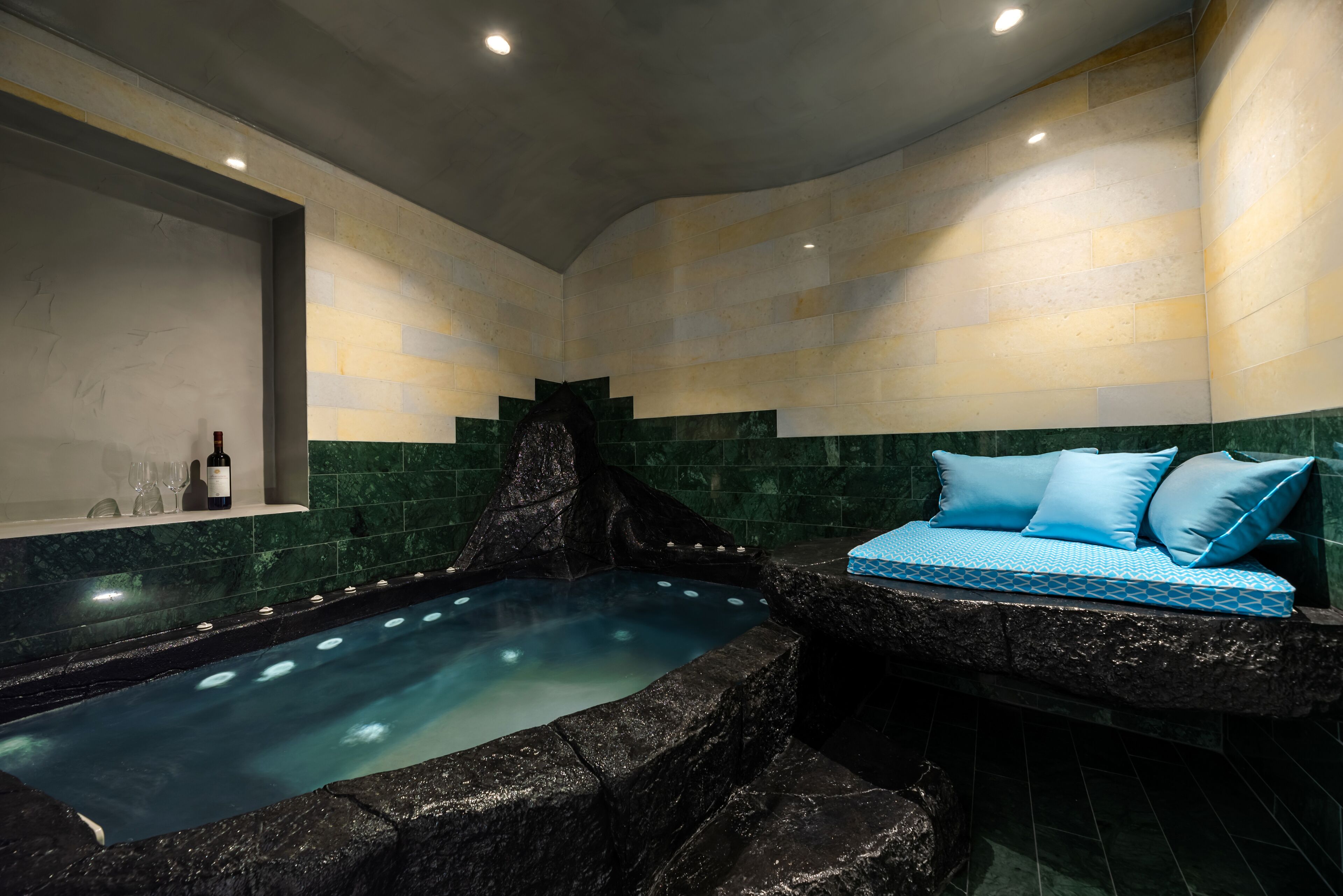 Un área de spa con un jacuzzi, una zona de estar con cojines azules y un estante con copas y botellas de vino.