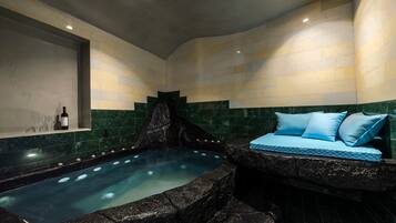 Un área de spa con un jacuzzi, una zona de estar con cojines azules y un estante con copas y botellas de vino.