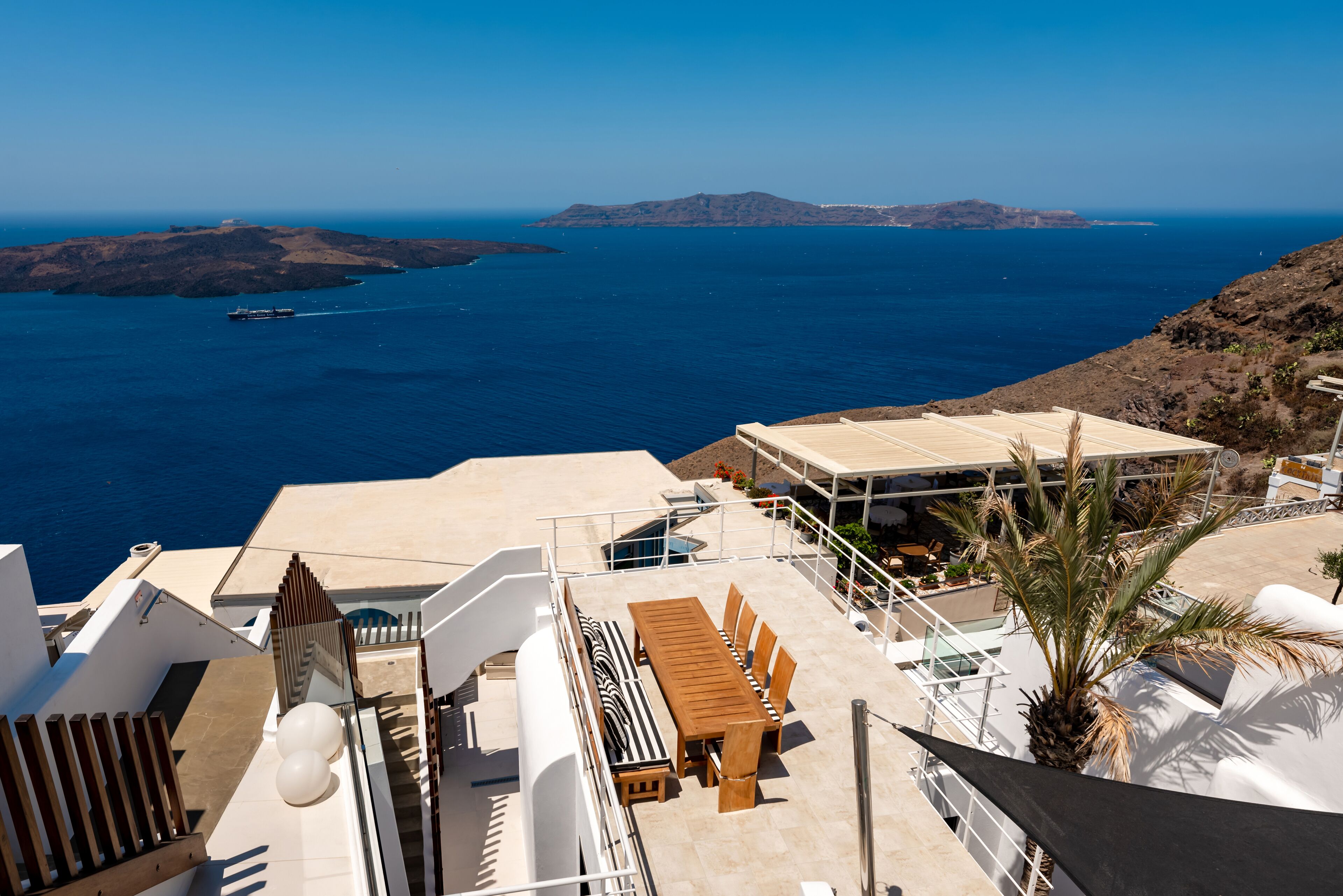 A rooftop terrace with a wooden table and chairs, overlooking a coastal landscape with clear blue waters and distant islands.