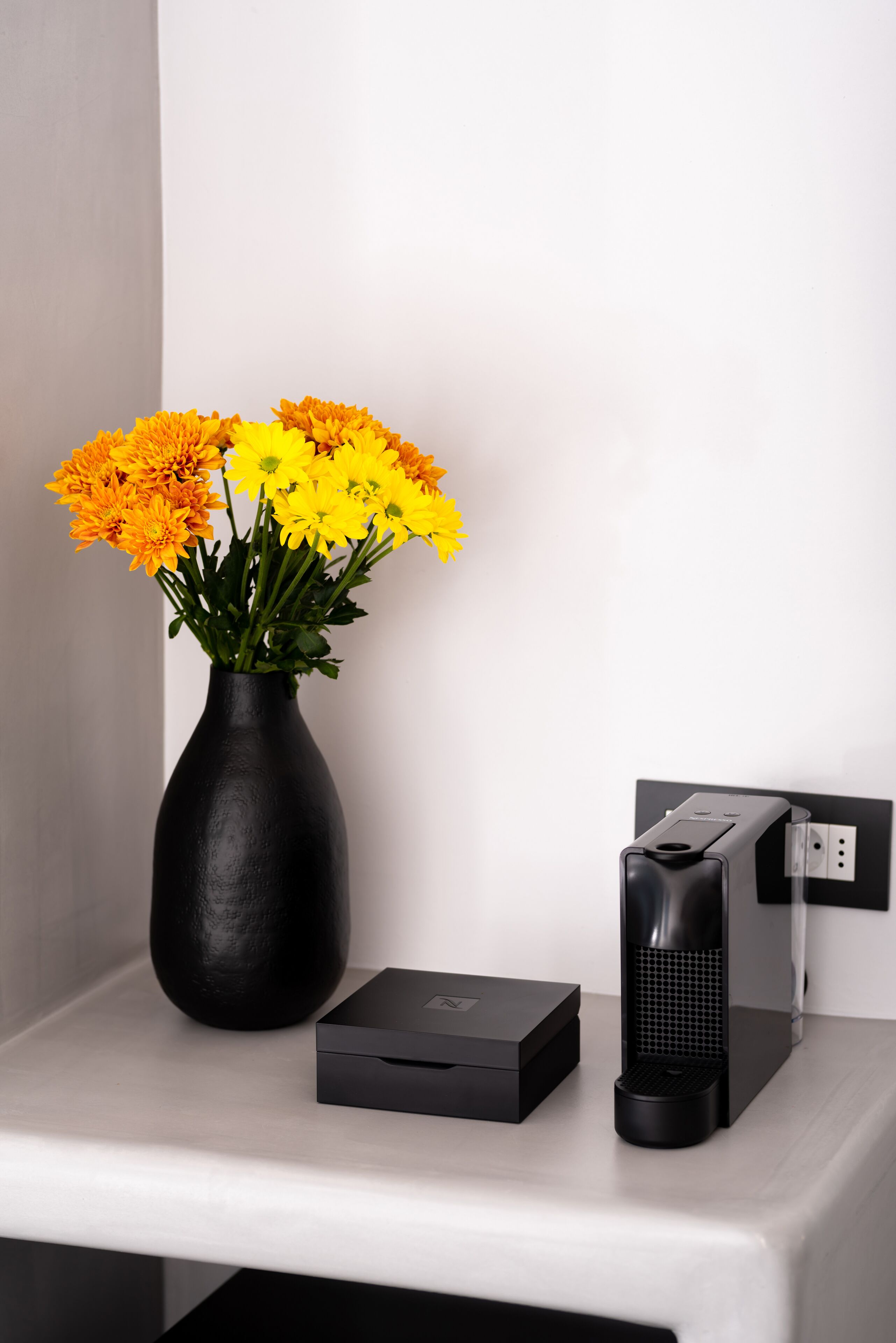 A black vase with yellow flowers on a white surface next to a black electronic device.