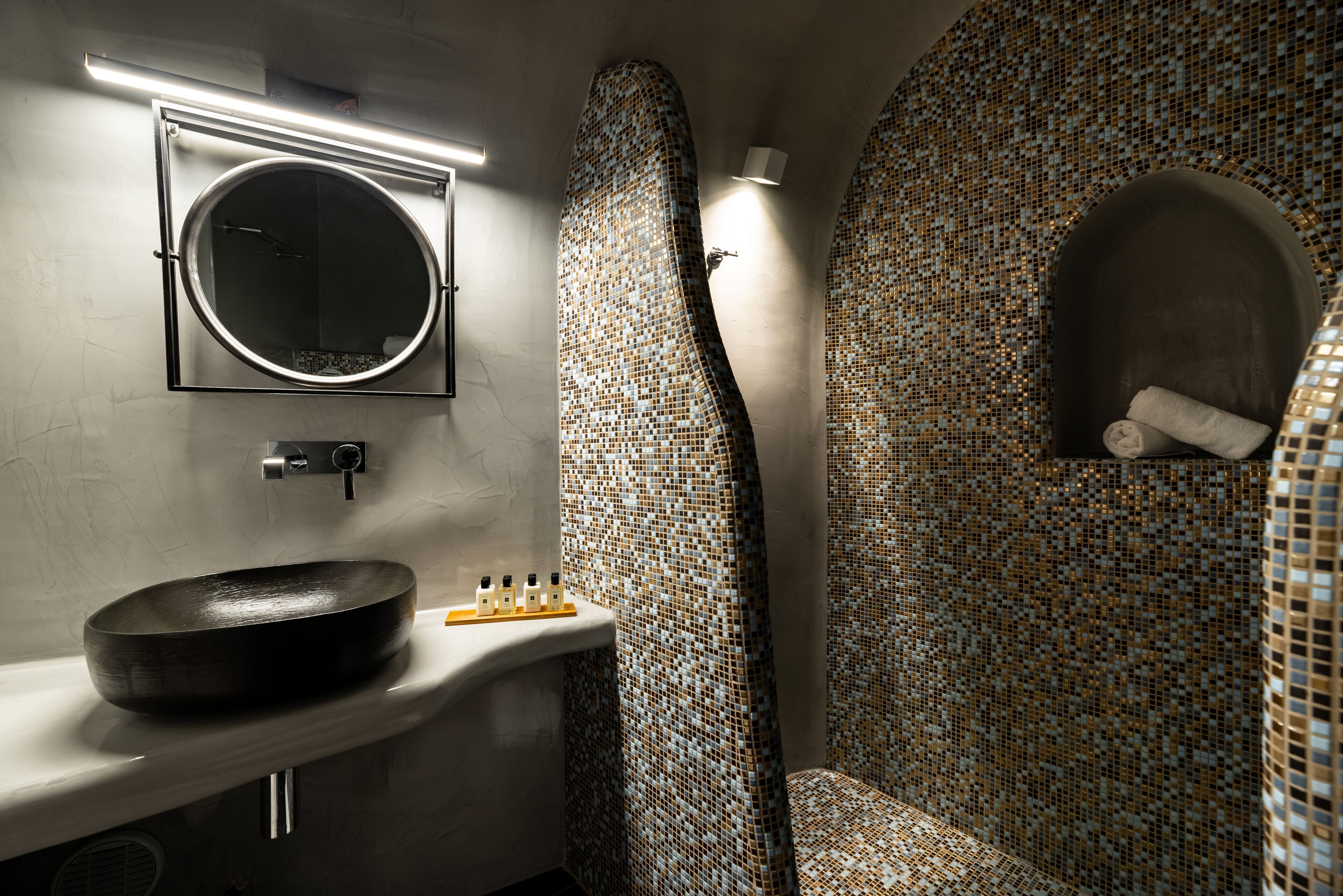 Un bany amb un mirall rodó, un lavabo i una dutxa amb rajoles de mosaic.