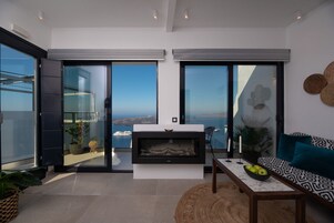 A modern living room with a fireplace, a sofa, a coffee table, and a view of the ocean through large sliding doors.