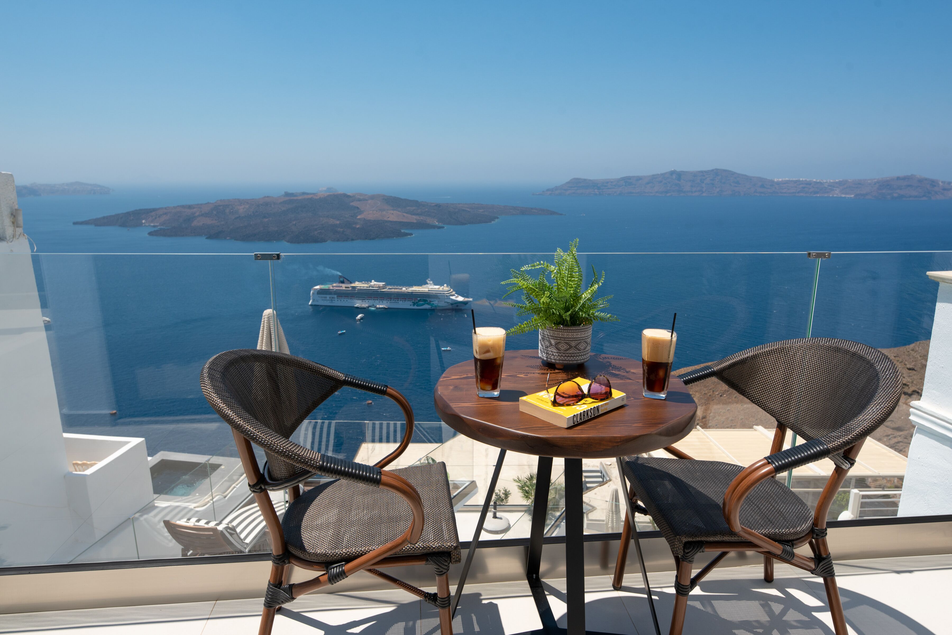 A balcony with wicker chairs, a table with drinks and a book, and a view of the sea and islands.