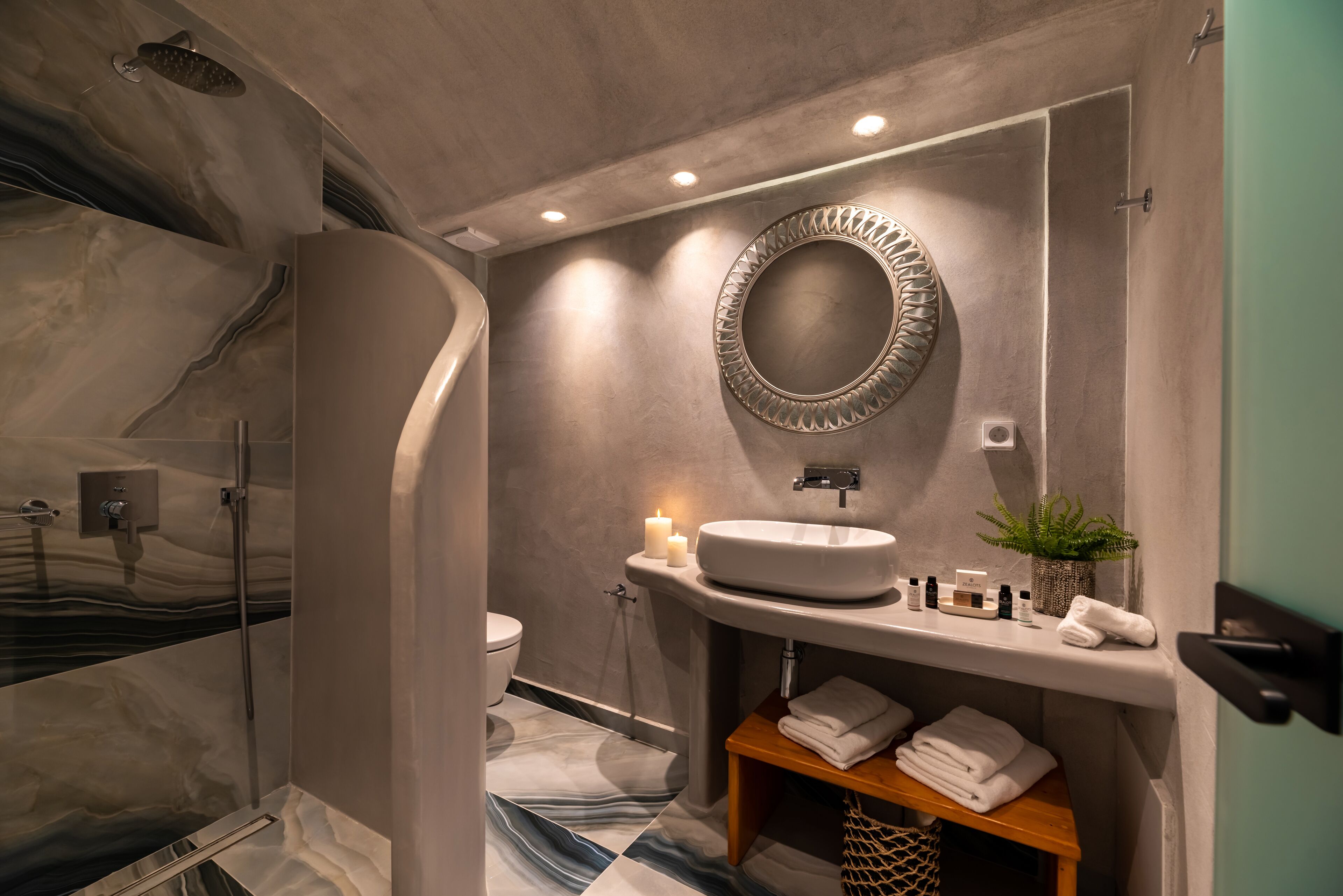 A bathroom with a marble floor, a sink with a mirror, a shower, and a shelf with towels.