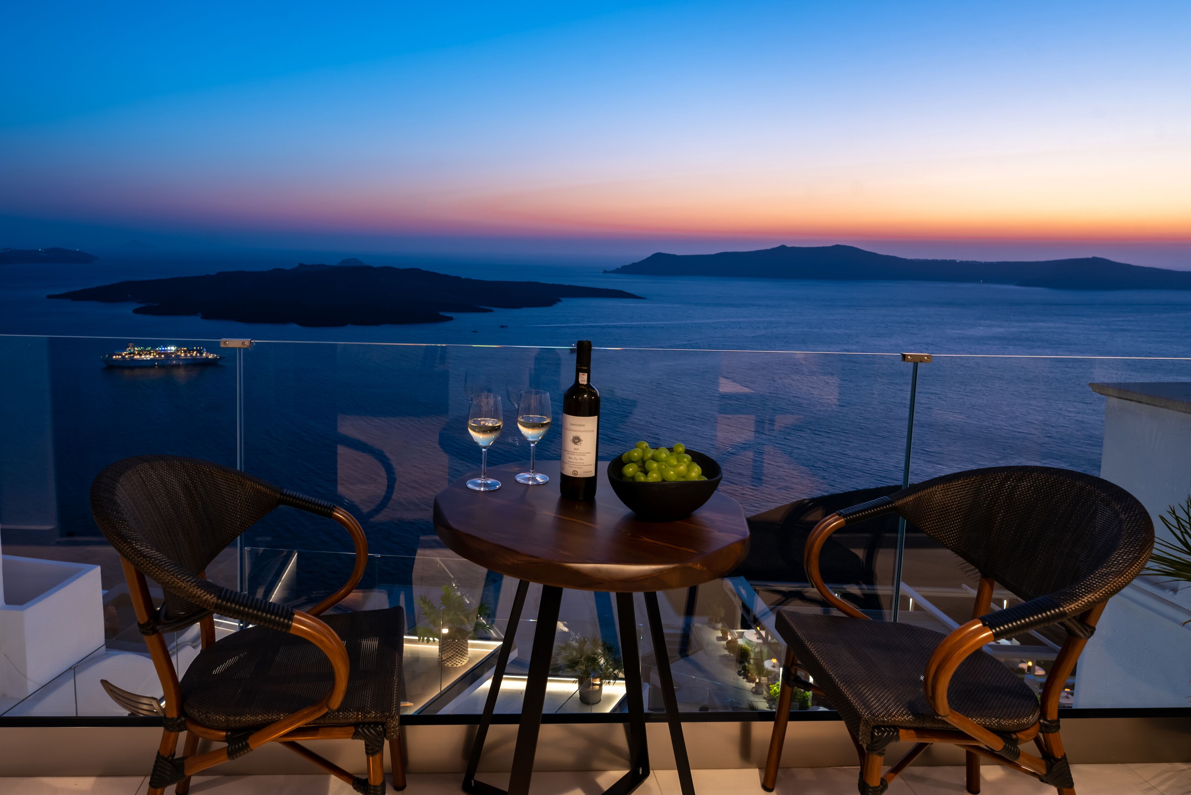 A balcony with two wicker chairs, a small round table with a bottle of wine, two glasses, and a bowl of fruit.