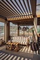 Un área de estar al aire libre iluminada por el sol con vista al mar, que incluye un banco de madera, un sofá acolchado y una mesa de centro.