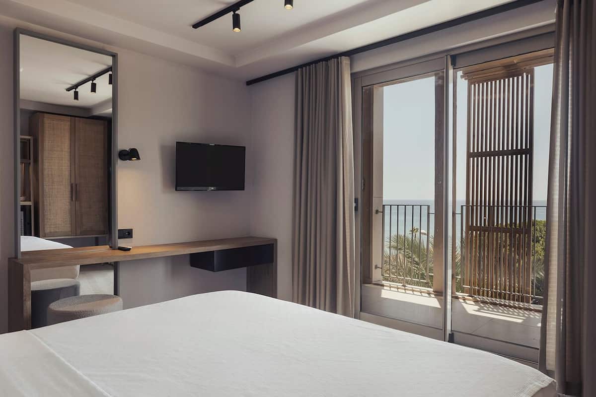 A modern hotel room with a large bed, a mounted TV, a bedside table, a mirror, and a balcony with a view of the sea.