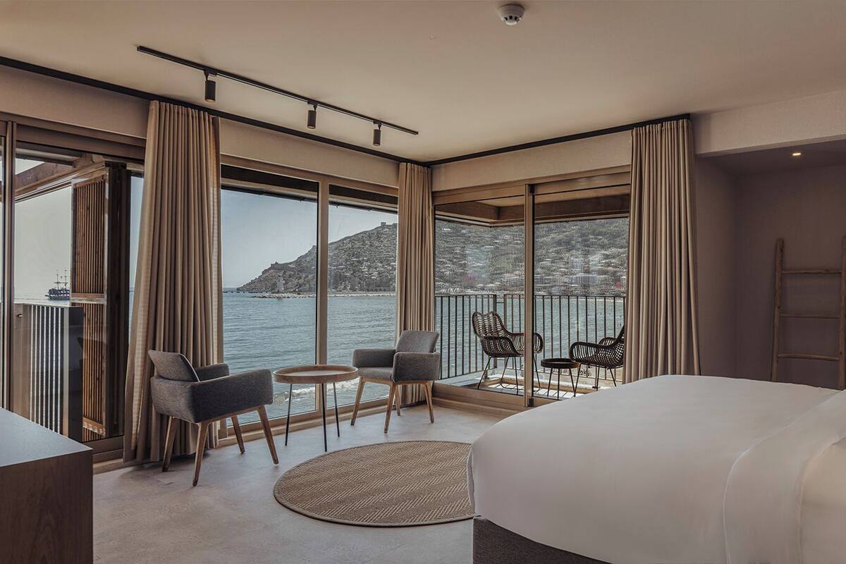 A modern hotel room with a large bed, a desk, a round table, and a balcony with chairs.