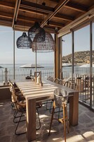 Un área de comedor al aire libre con mesa y sillas de madera, ventanas amplias y lámparas colgantes de mimbre.