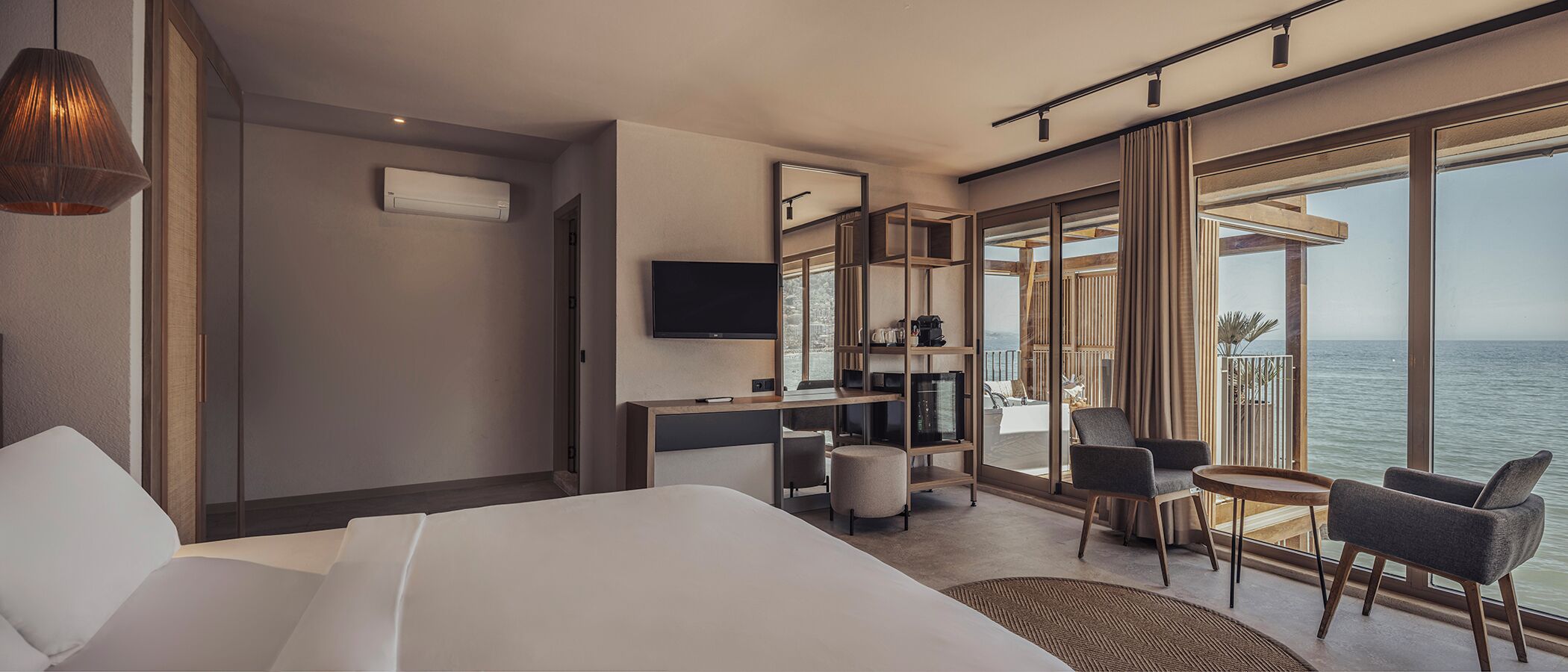 Una habitación de hotel moderna con una cama grande, un escritorio con televisor de pantalla plana, una zona de estar con sillas y un balcón con vista al mar.