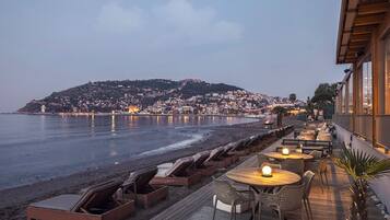 Terraza frente al mar con sillones y mesas, con vistas a una playa y un pueblo.