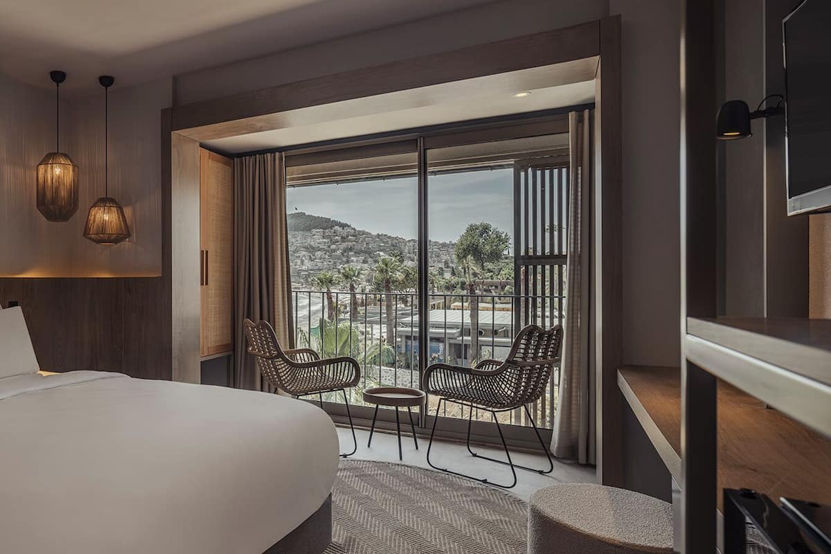 A modern hotel room with a large bed, a balcony with wicker chairs, and a city view.