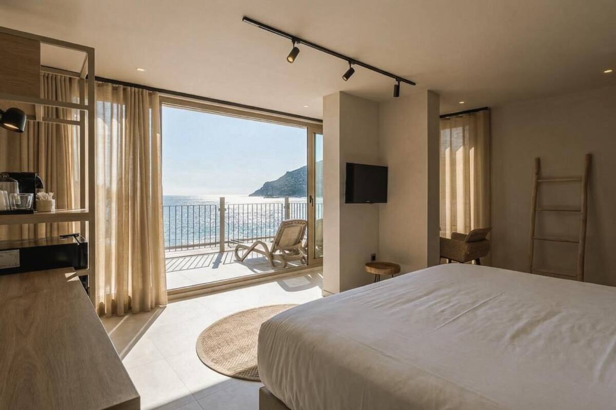 A hotel room with a large bed, a desk, a chair, a TV, and a balcony with a view of the sea.