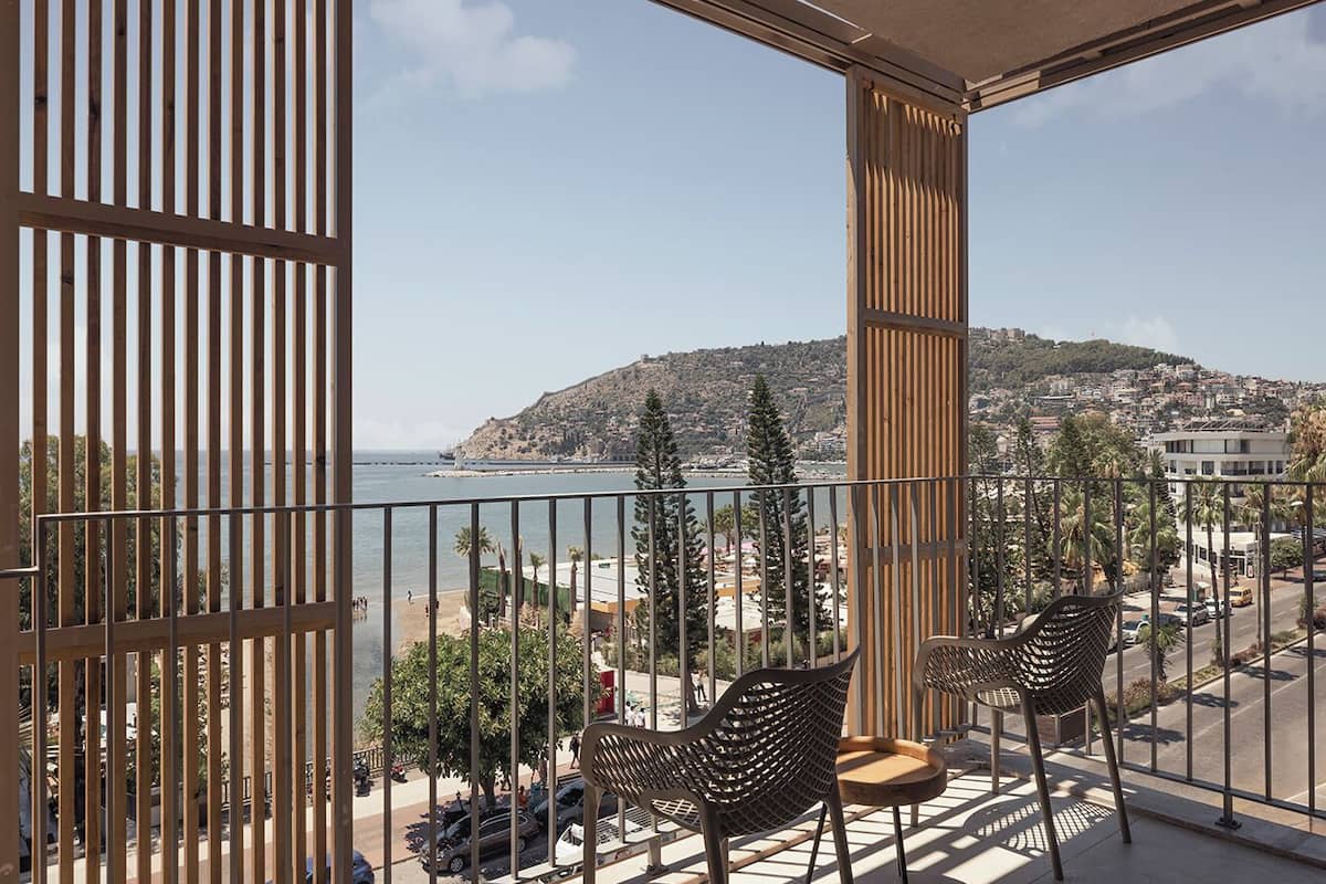 A balcony with two chairs and a table, overlooking a coastal cityscape with a hill and a body of water.