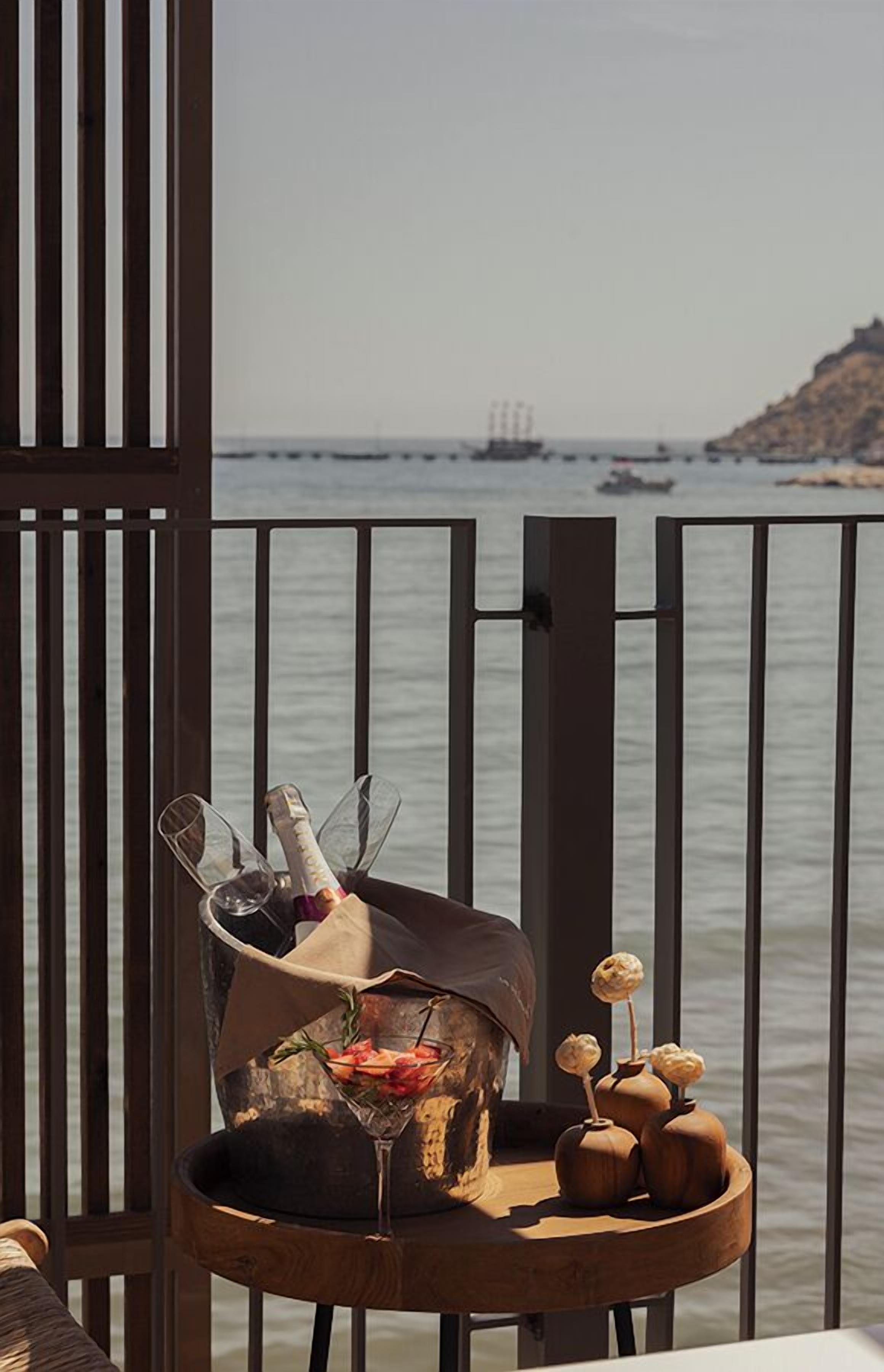 A wooden table with a bottle of champagne, a glass, and decorative vases by a railing overlooking a body of water.